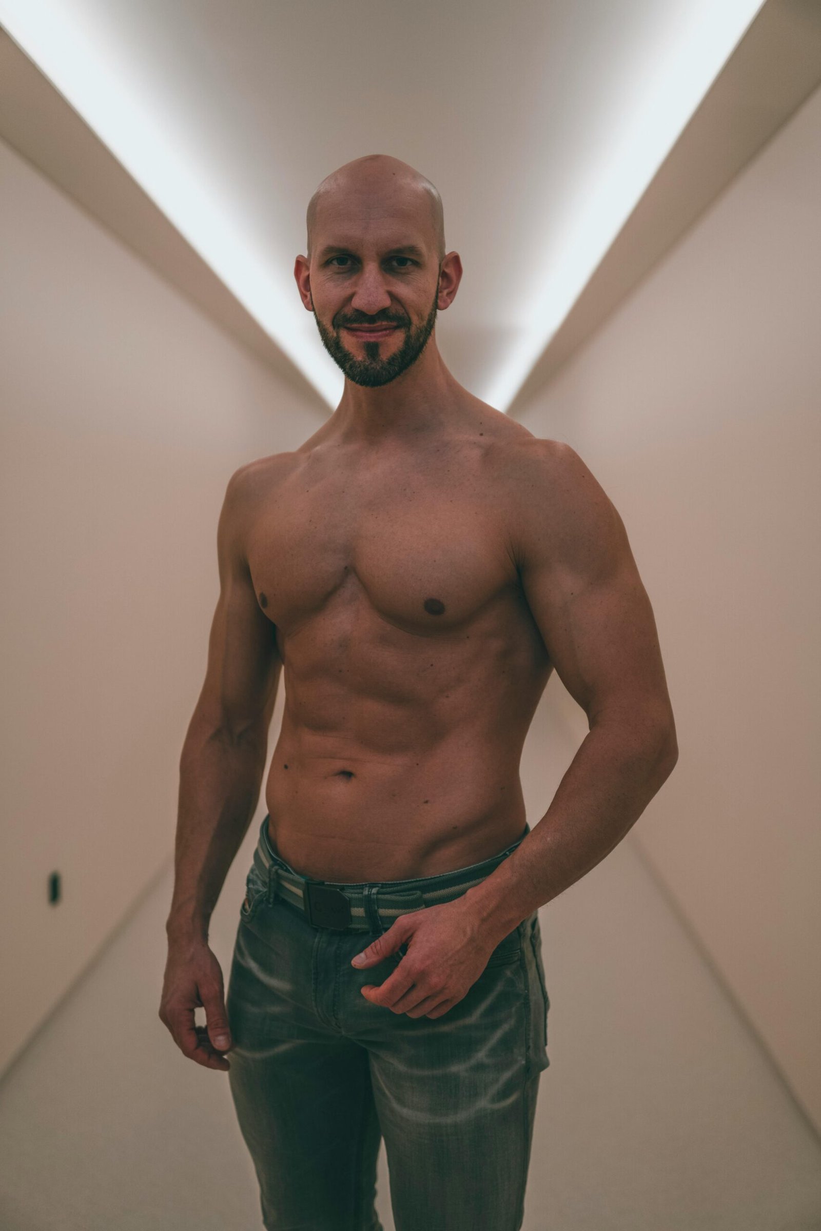 Shirtless bald man with beard standing confidently in minimalist hallway.