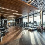Modern exercise bikes and treadmills and various fitness equipment in spacious gym with wooden floor and panoramic windows in sunlight